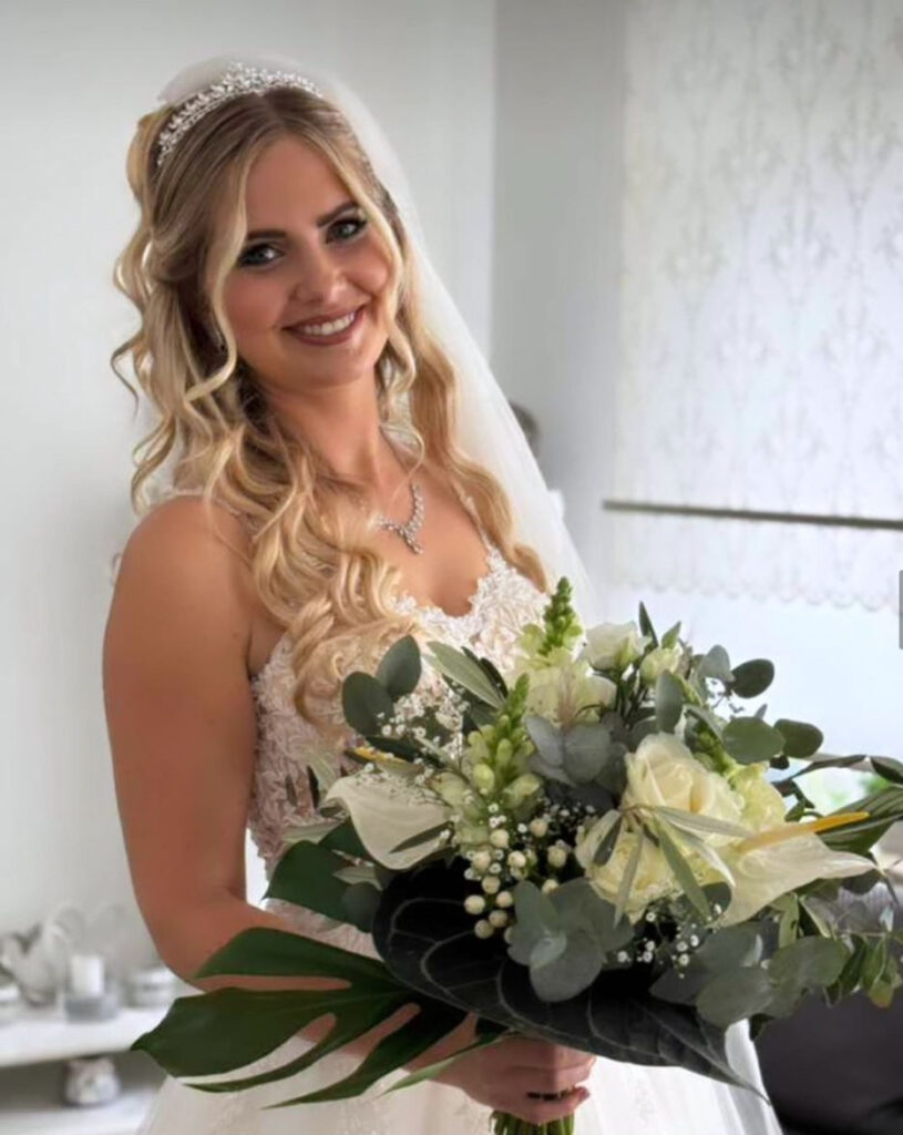 Eine lächelnde blonde Frau im Brautkleid hält einen großen Strauß weißer Blumen